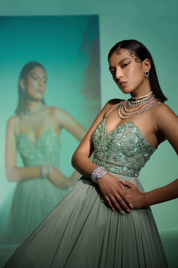 A woman modeling a floor-length sage green georgette anarkali gown with chic noodle strap sleeves, wearing jewelry and heels.